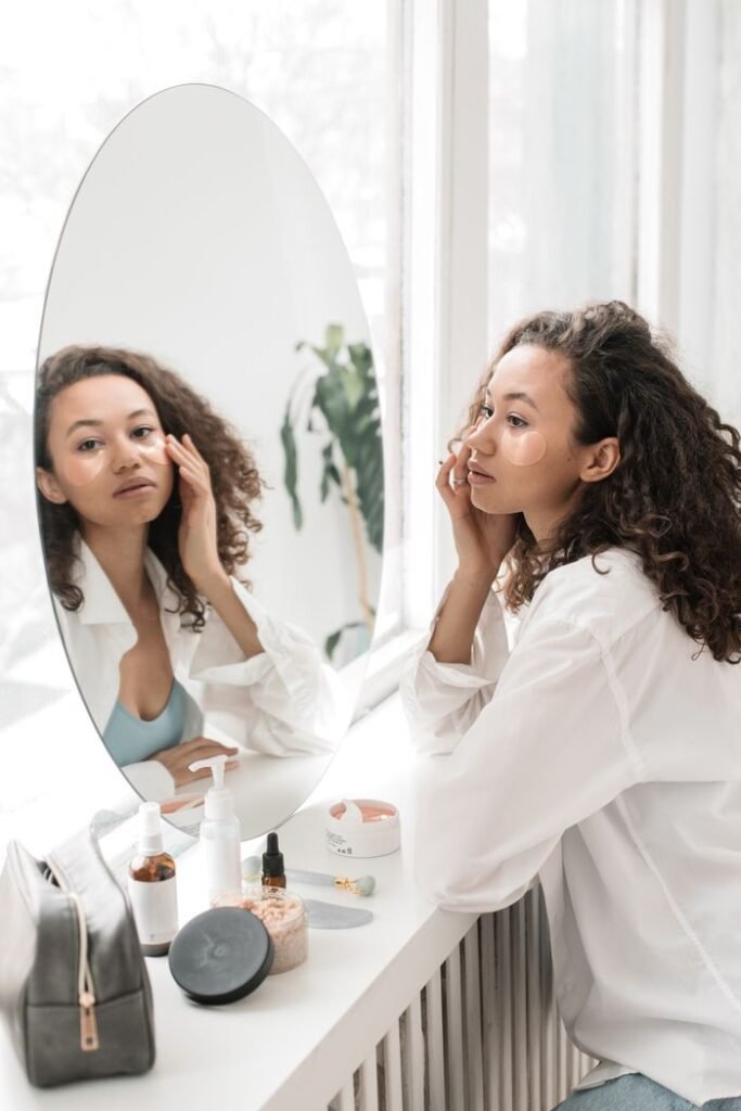 A woman applies skincare products, sitting at a vanity with a mirror in a bright room.